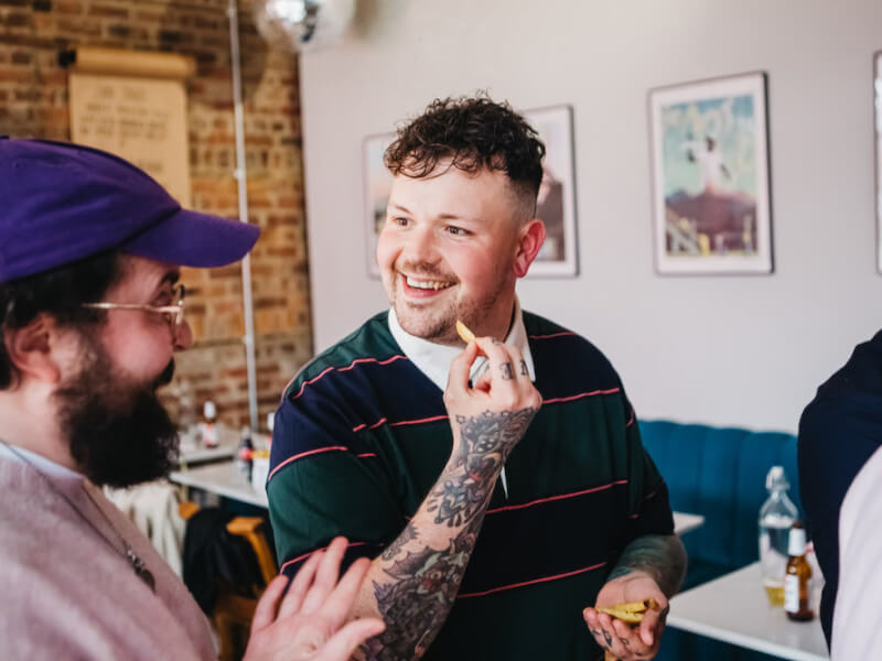 Man smiling at friend in a purple cap whilst tasting chips and dips at a pizza party.
